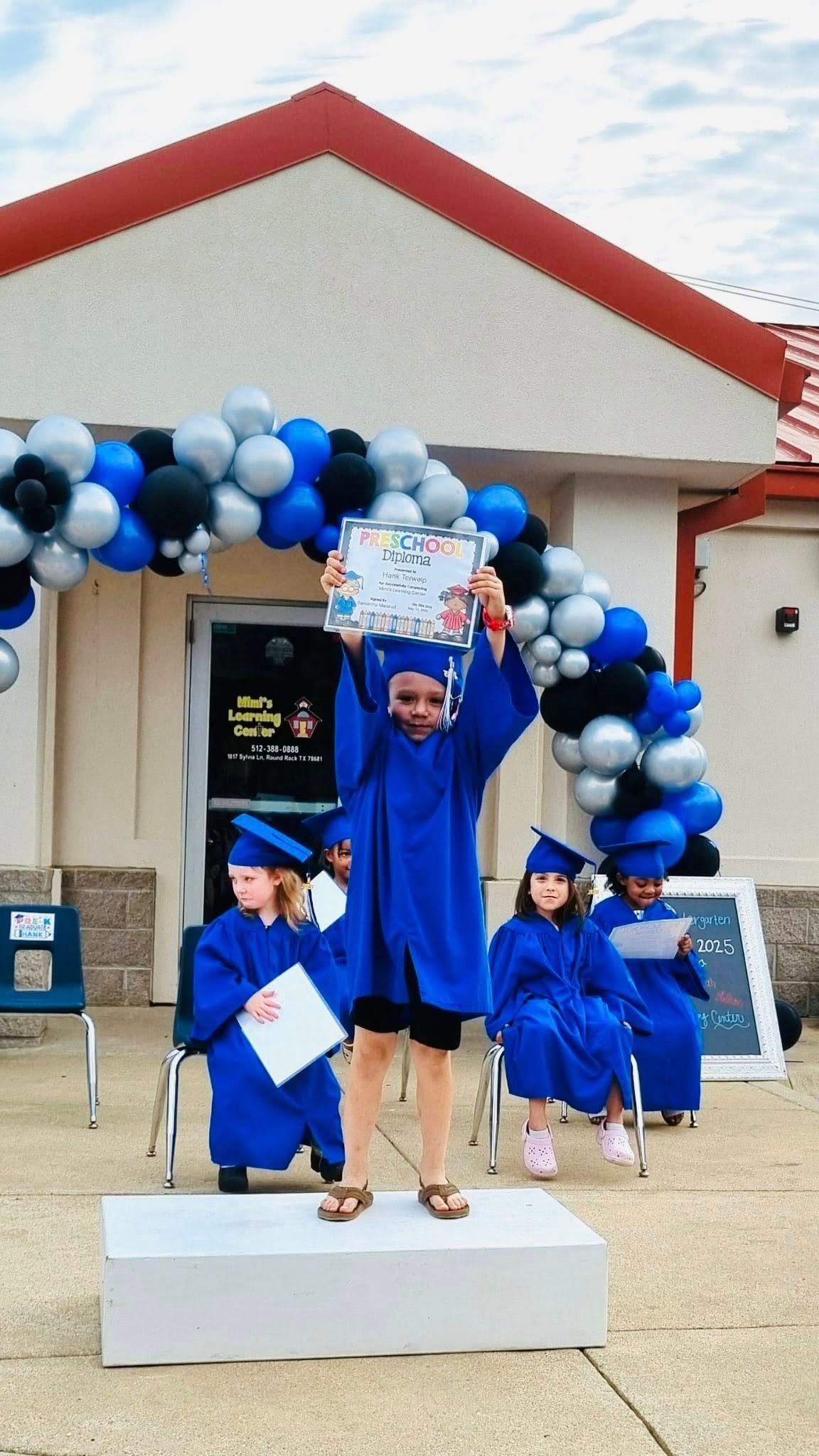 Three graduates in blue caps and gowns pose on a podium under a black, white, and blue balloon arch.