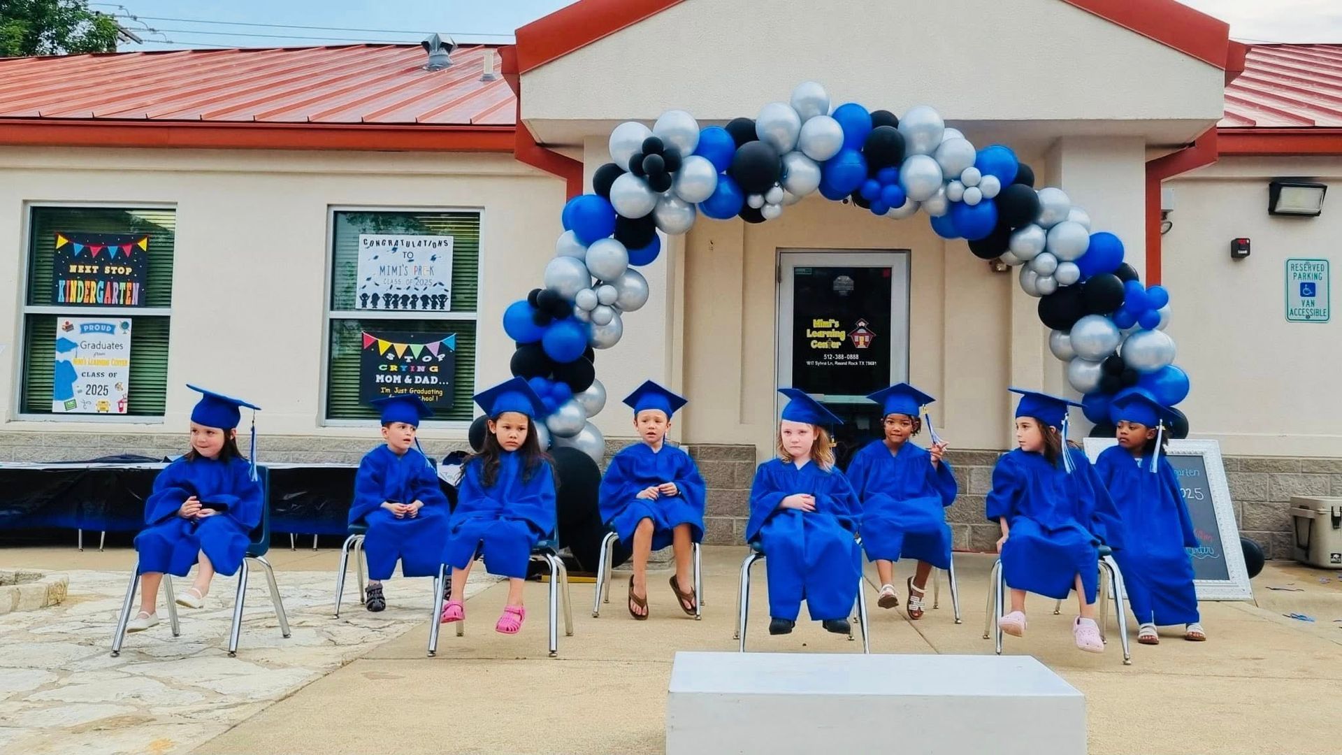 Graduation ceremony with children in blue caps and gowns seated under black, white, and blue balloon arch