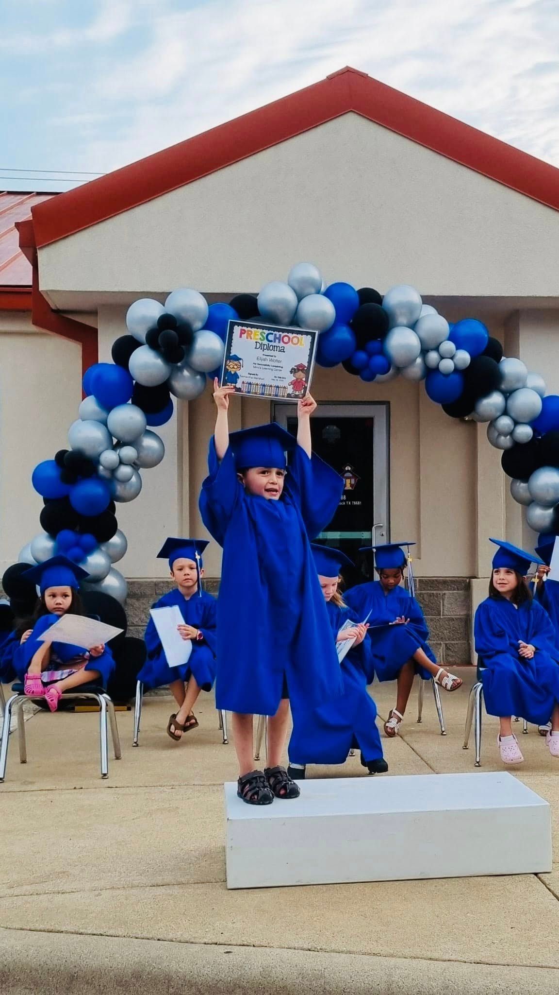 Child in blue graduation gown raises diploma on stage under black, white, and blue balloon arch