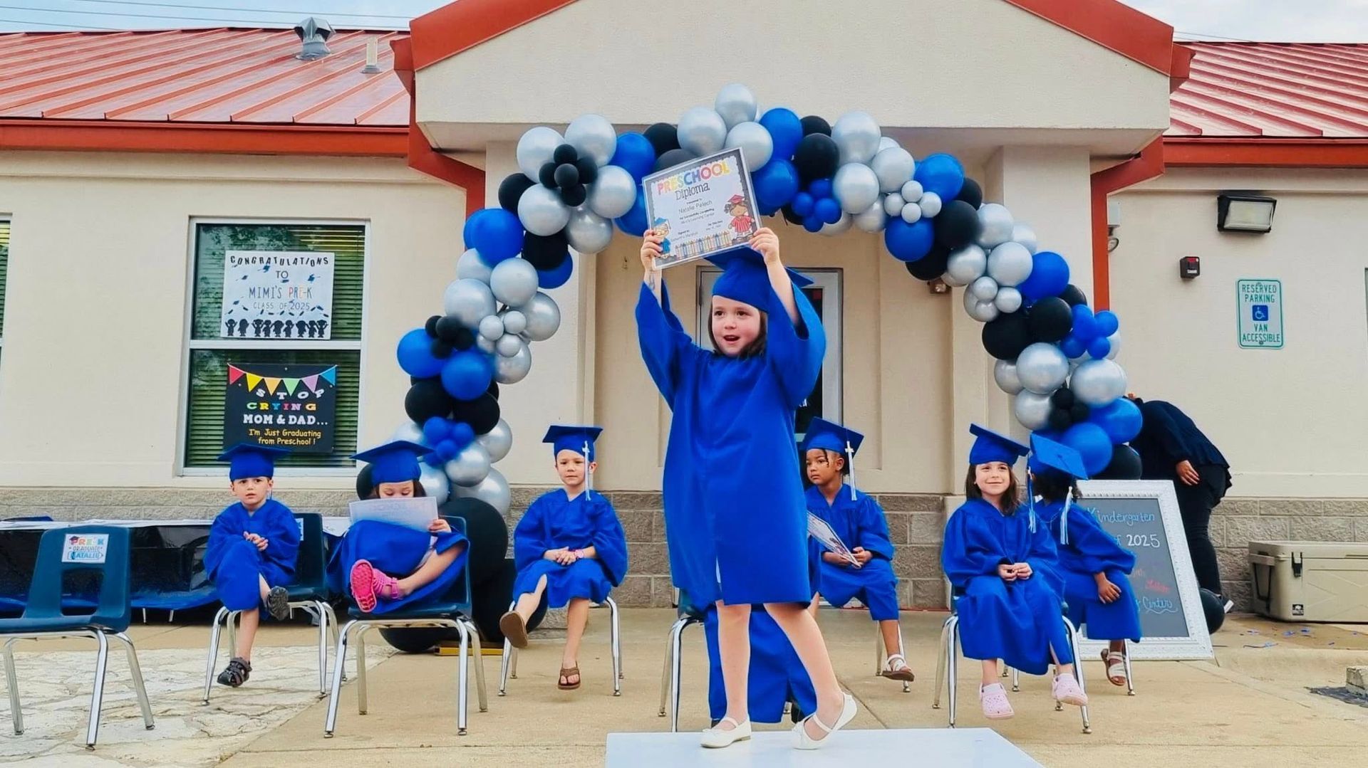 Children in blue uniforms pose on stage under a blue-and-white balloon arch at a school event