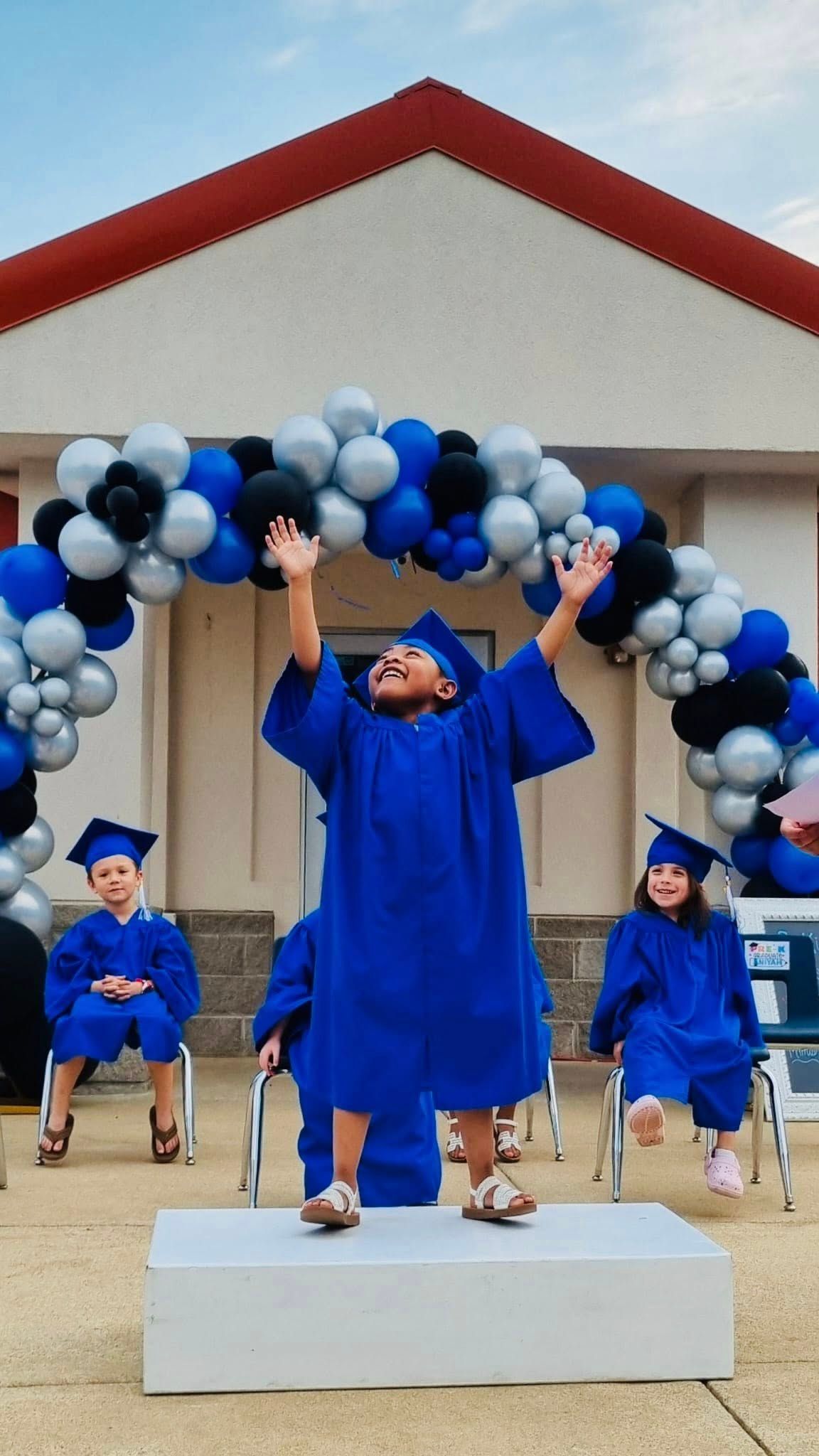Graduates in blue caps and gowns celebrate onstage beneath a black-and-white balloon arch