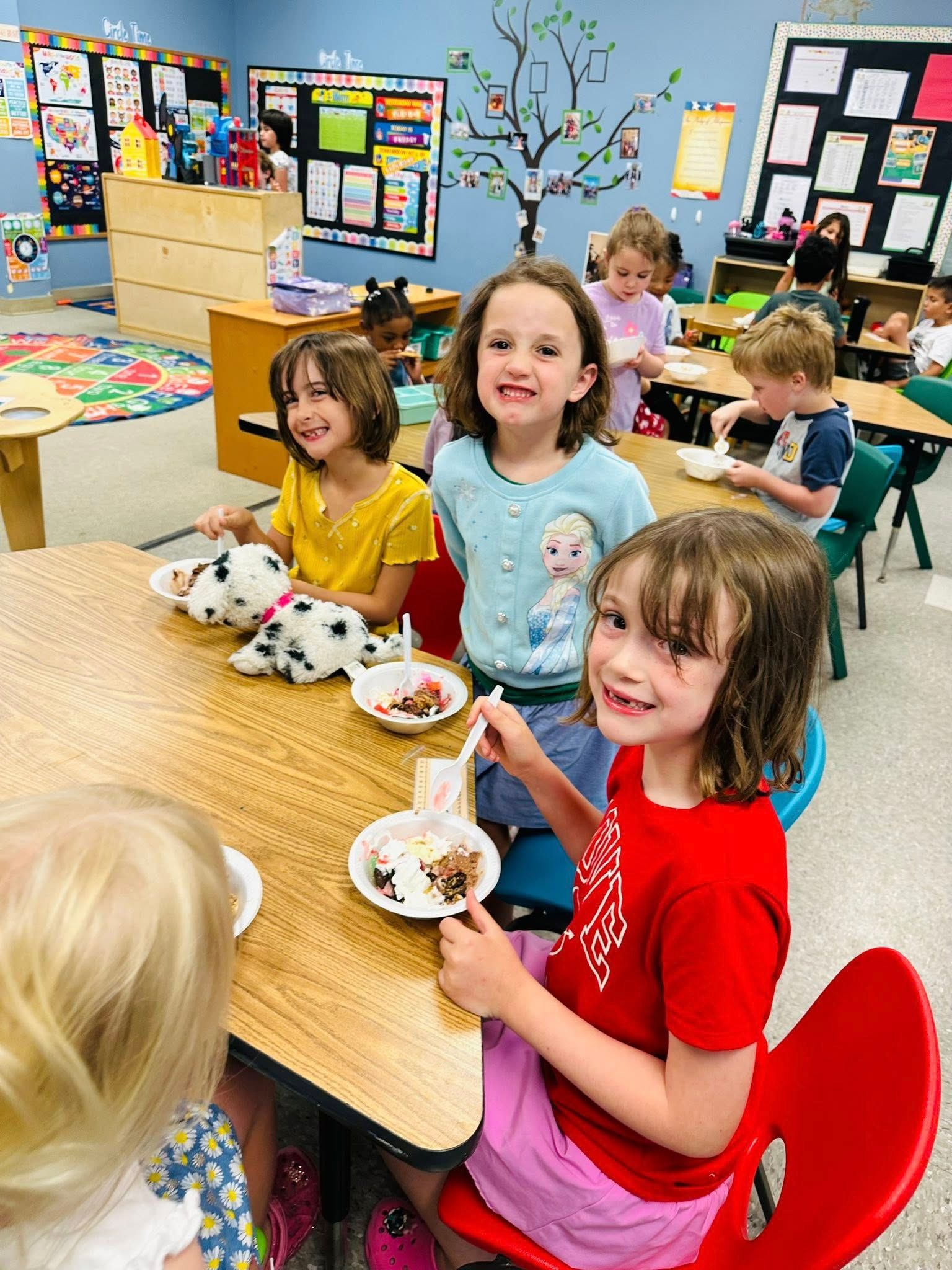 Children seated at a classroom table with tea sets, smiling in a colorful play area.
