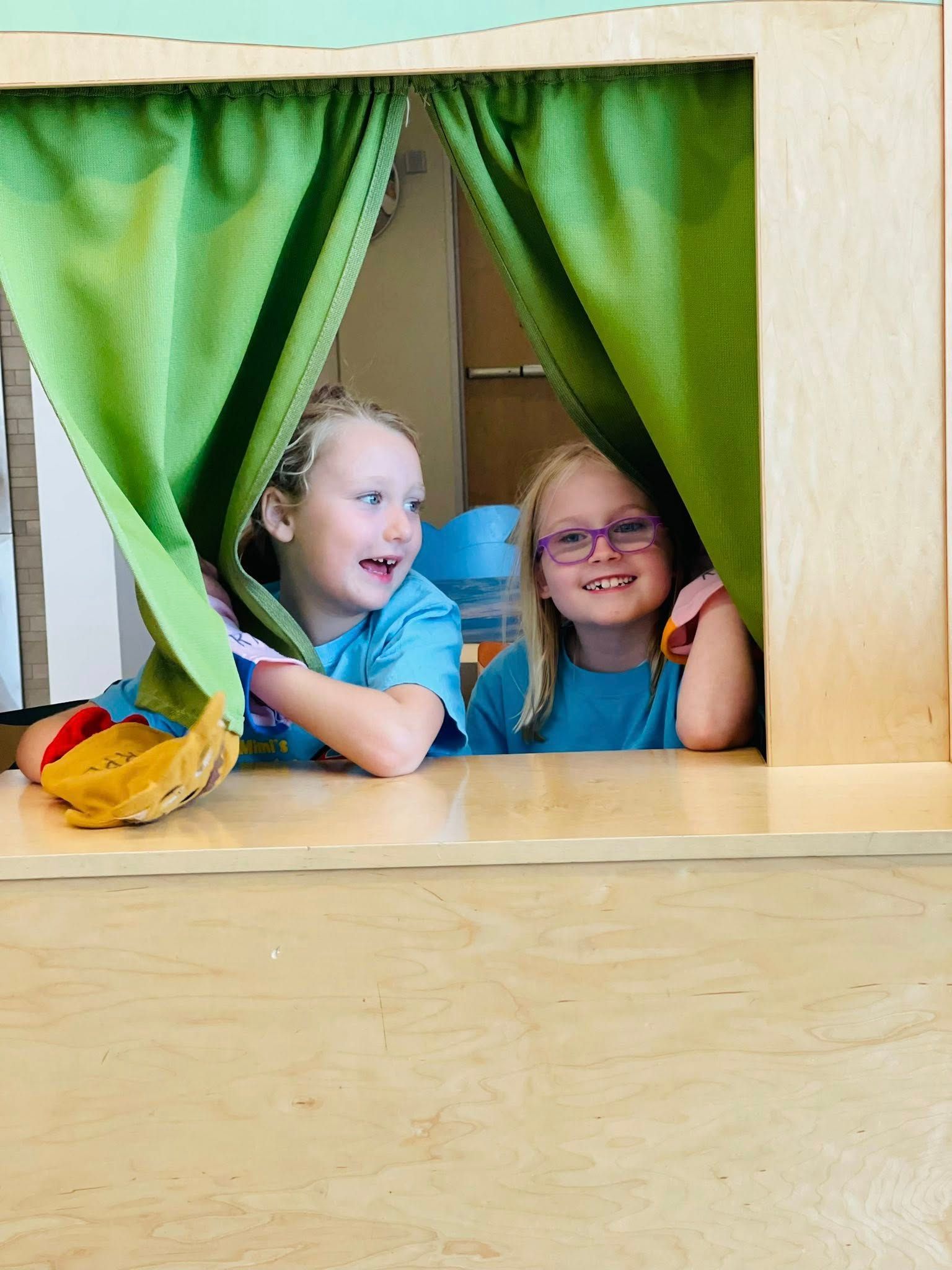 Two children peeking through green curtains in a wooden playhouse, smiling together