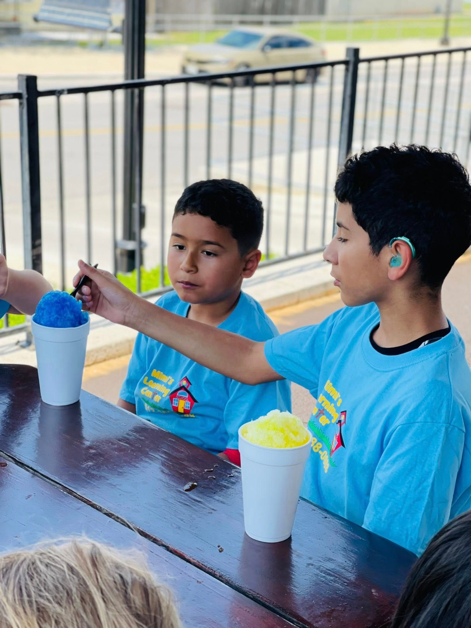Two children in blue shirts sitting at a picnic table with shaved ice cups outdoors.