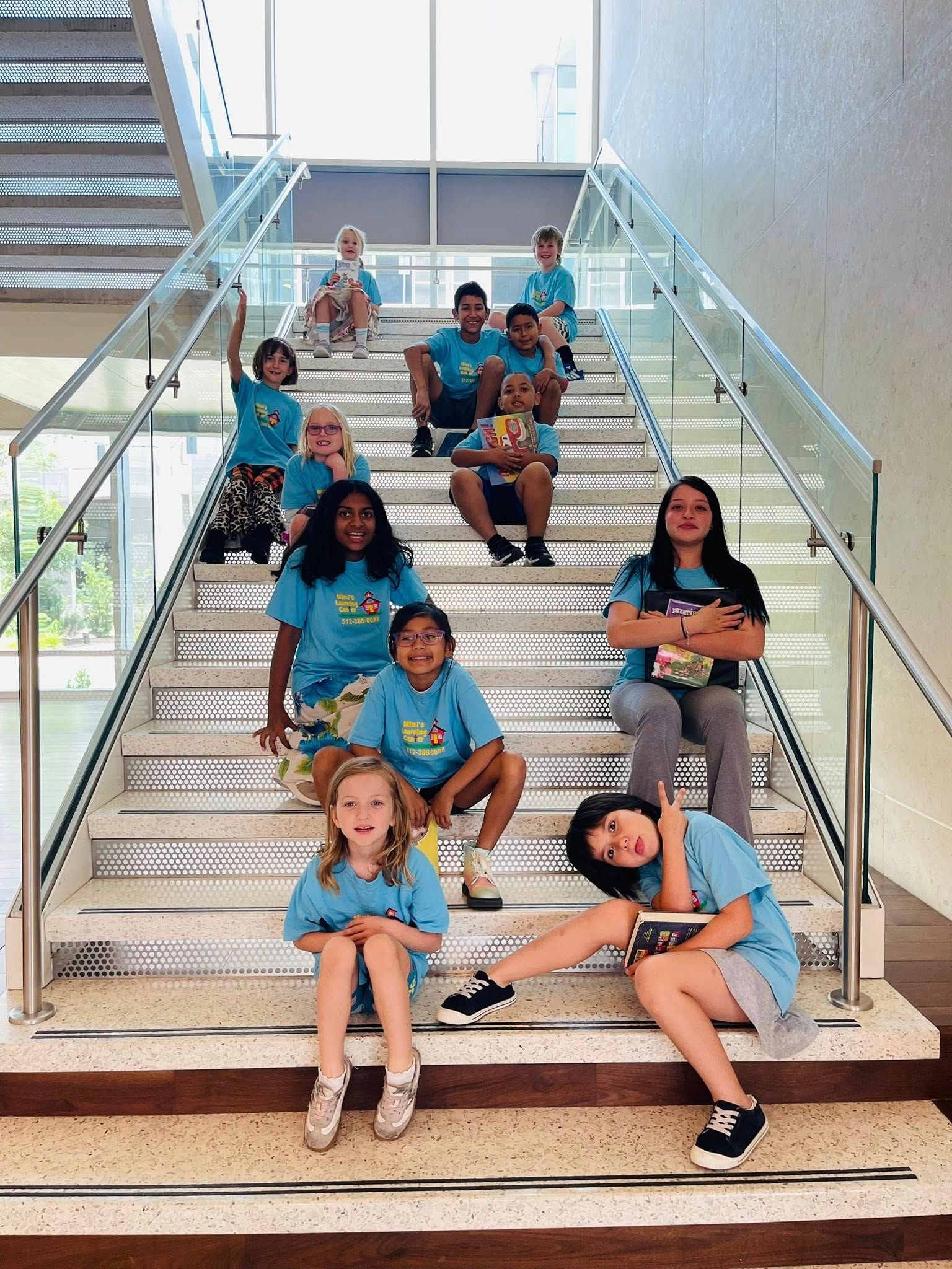 Students sitting on stairs in a bright indoor atrium, holding books and notebooks.