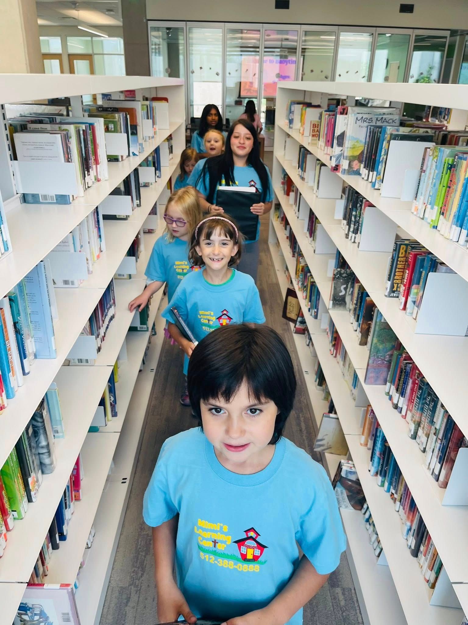 Children in blue shirts standing in a narrow library aisle between tall bookshelves