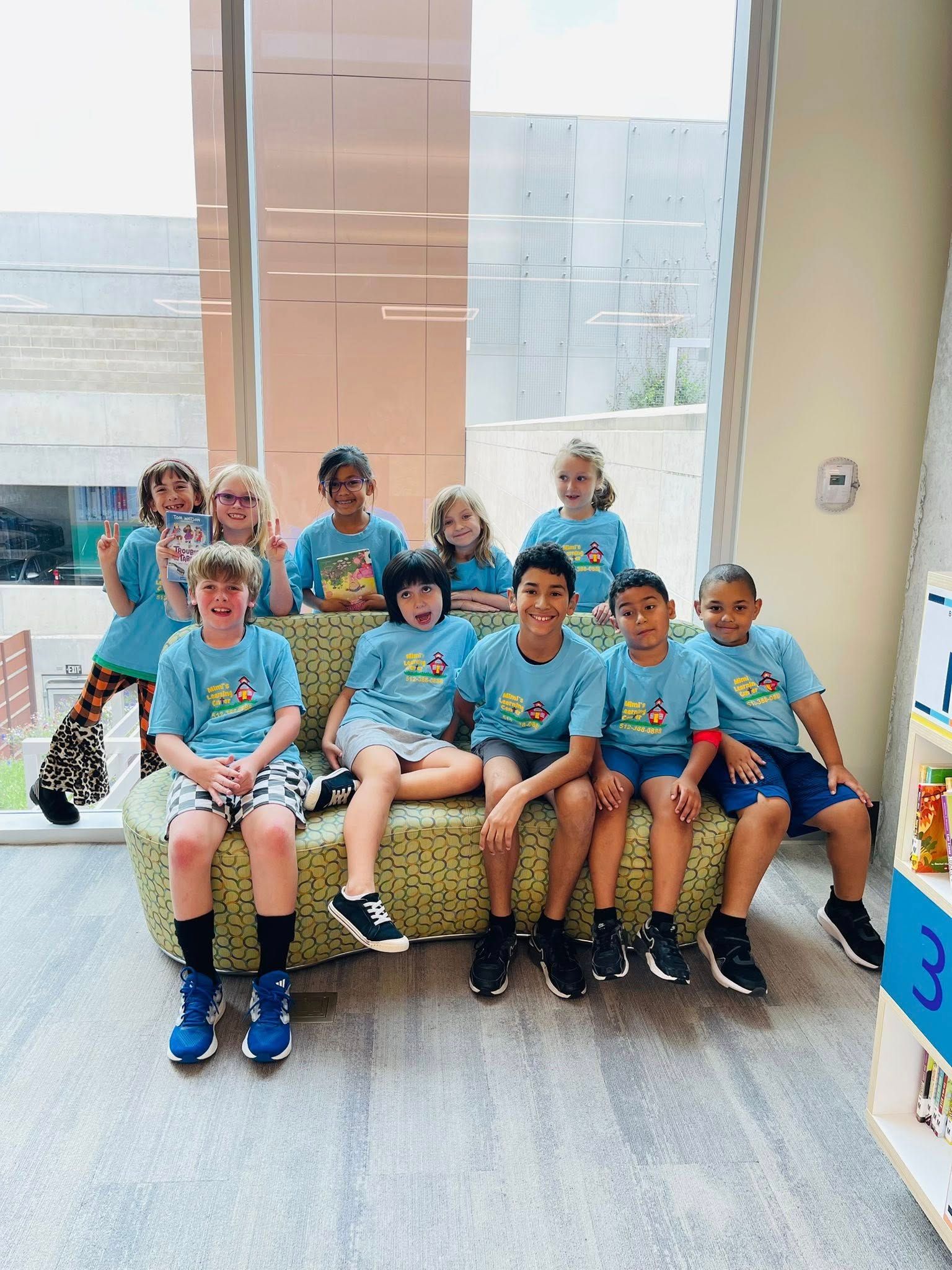 Group of children in blue shirts sitting on a bench in a bright indoor lobby, smiling at the camera.