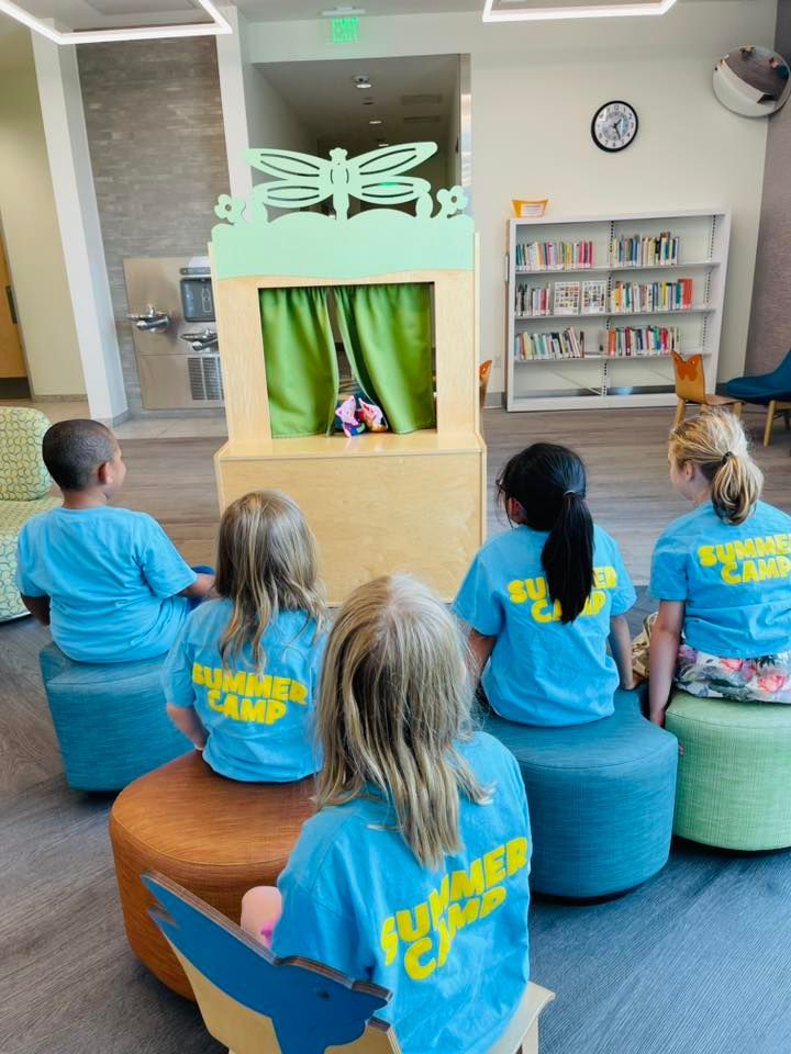 Children in blue shirts watch a puppet show in a bright library room