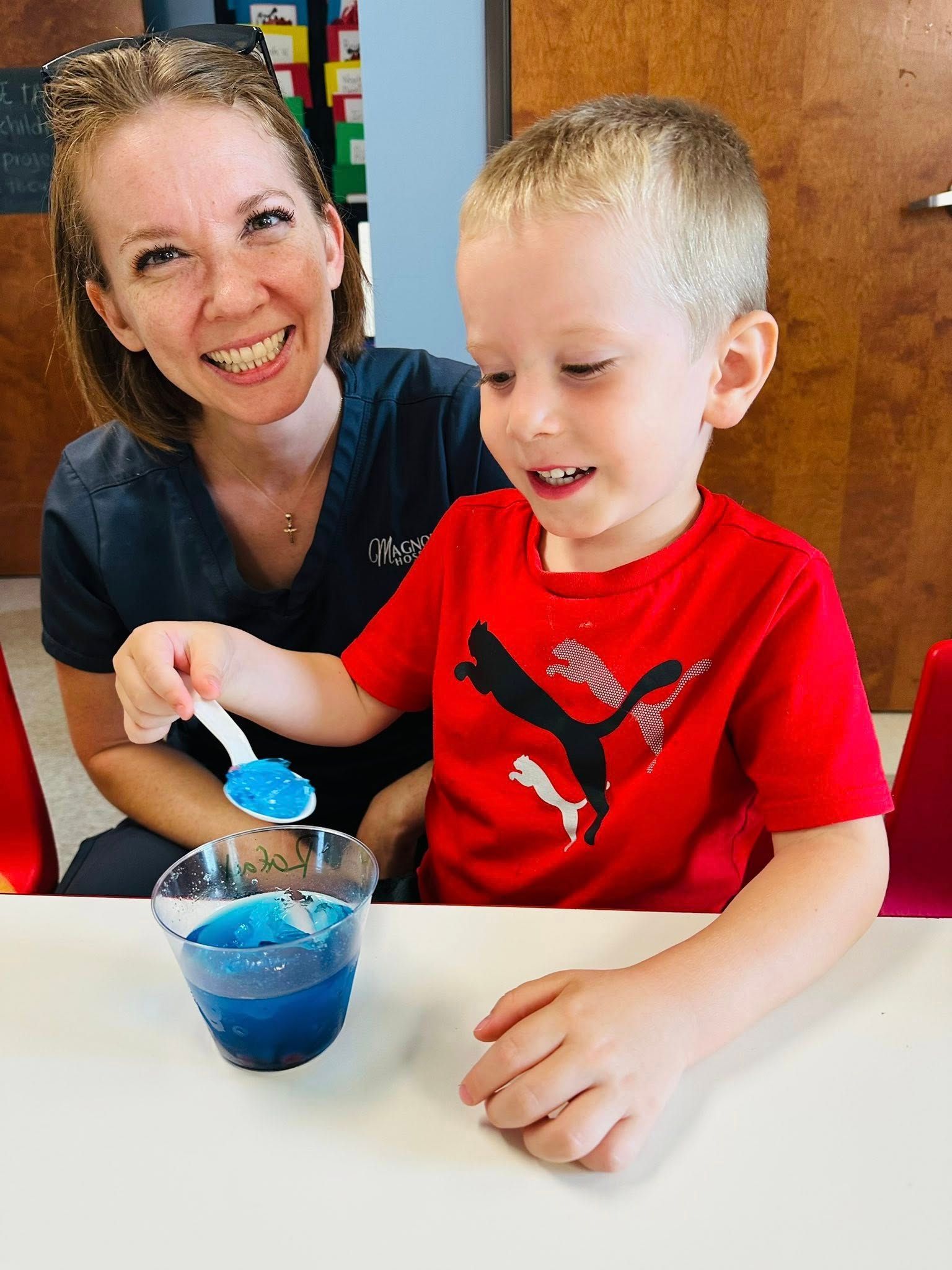 Smiling adult helping a child stir blue liquid in a cup at a table
