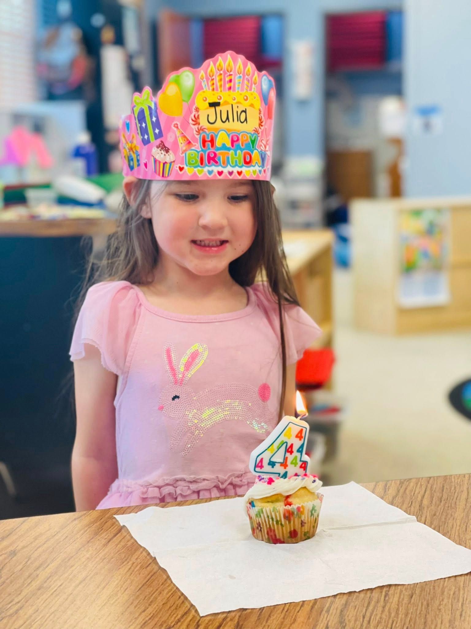 Child in a pink shirt wearing a colorful birthday crown, smiling at a cupcake on a table.