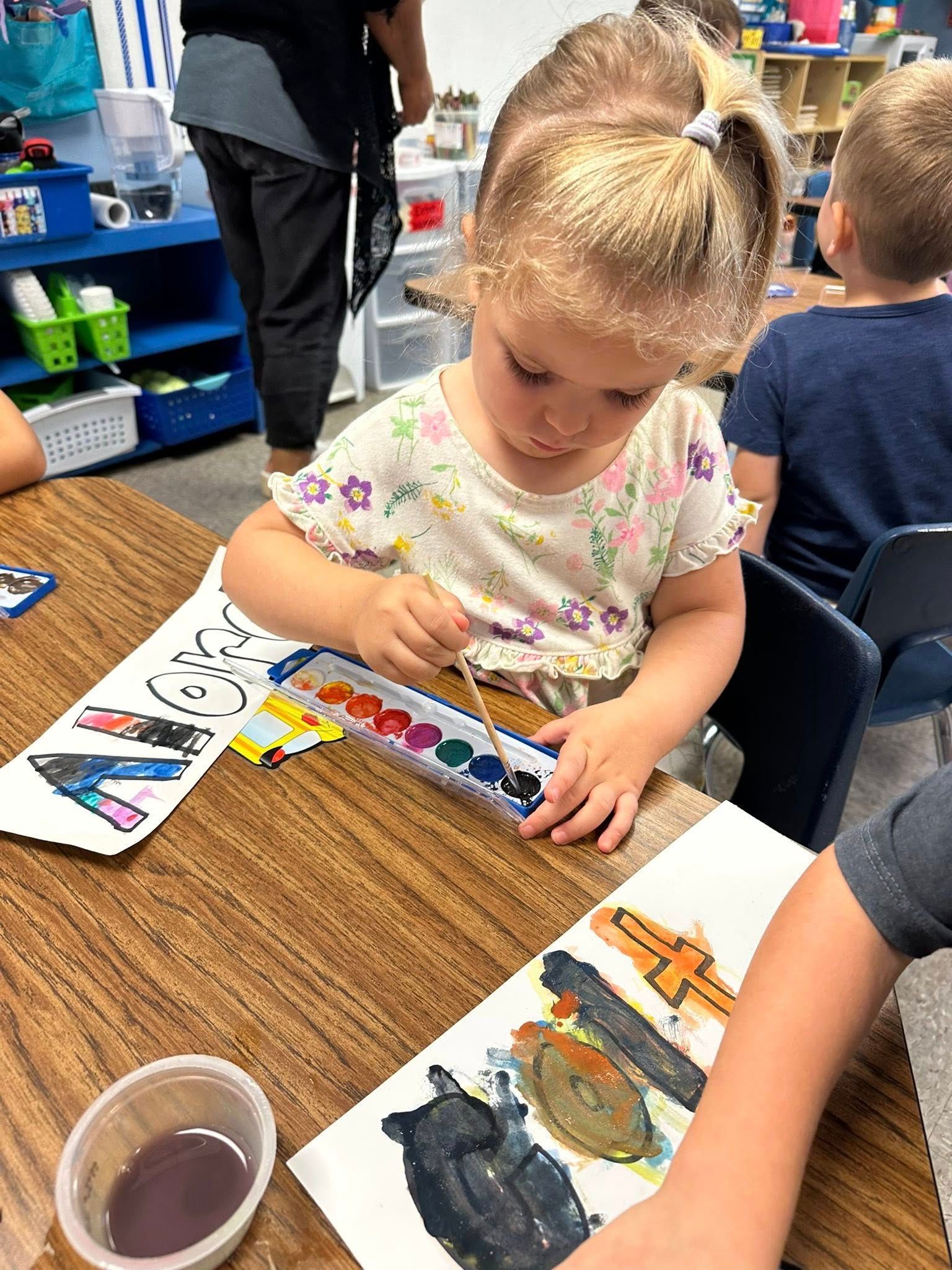 Child painting at a classroom table with watercolors and a cup of purple water.