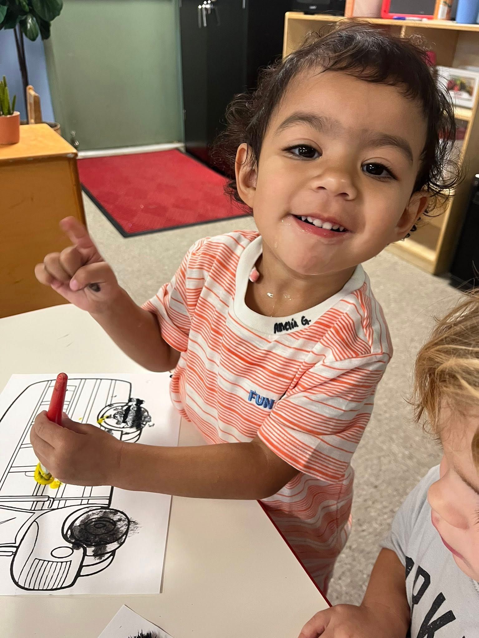 Smiling child in striped shirt pointing at a coloring page with crayons at a table