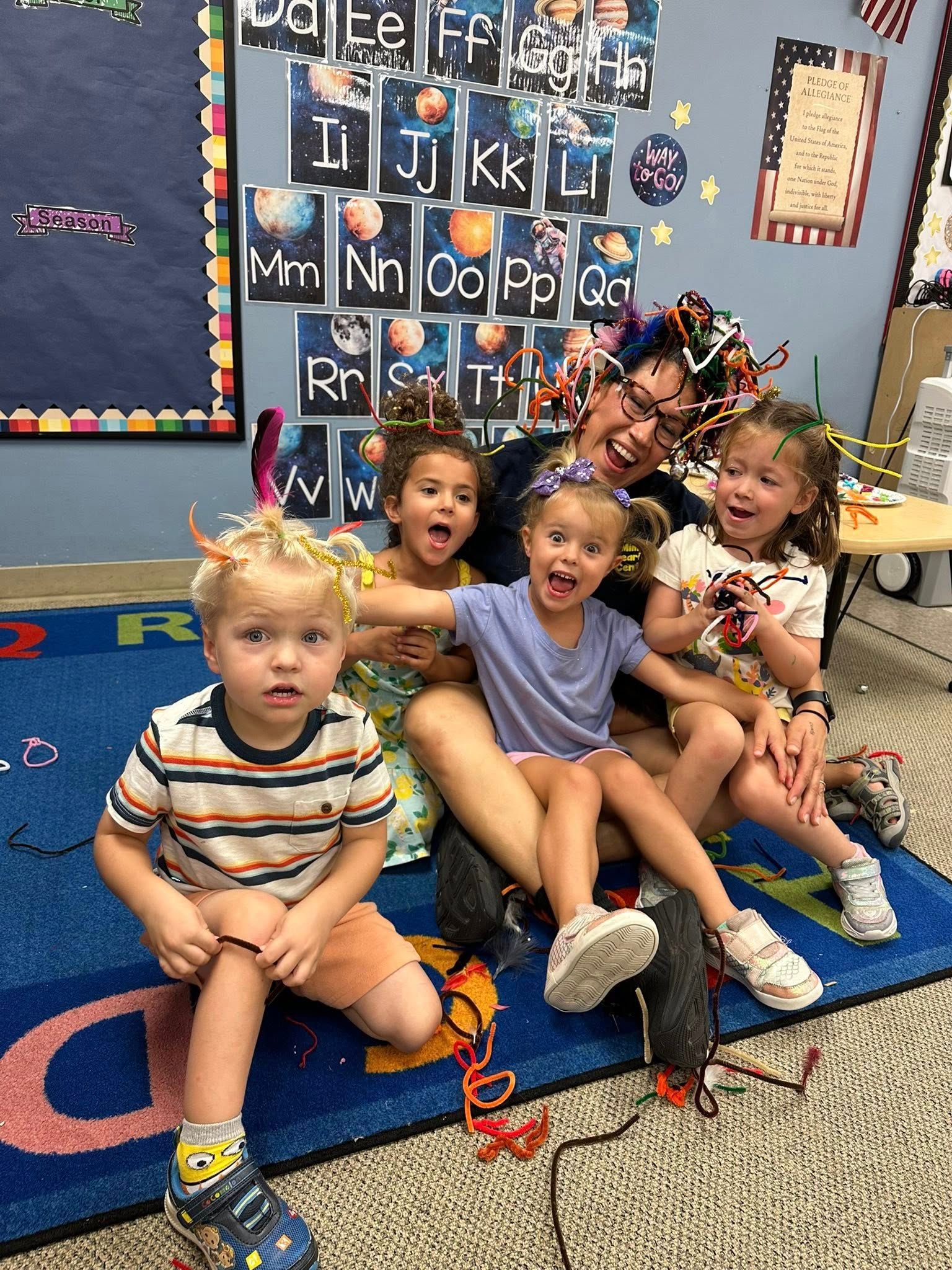 Children sitting on a classroom rug, smiling in front of an alphabet wall display.