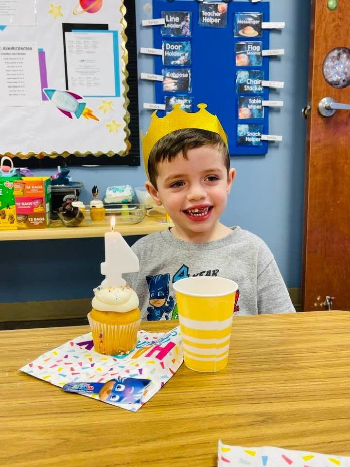 Child smiling at a classroom table with a cupcake and striped cup, with blue bulletin boards behind them
