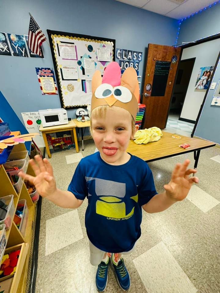 Child wearing a paper turkey hat and blue shirt, smiling in a classroom with tables and drawings behind them