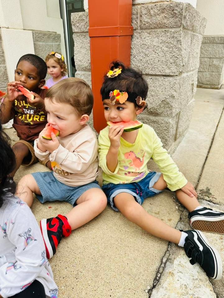 Three children sit on a sidewalk eating ice pops outside a stone building.