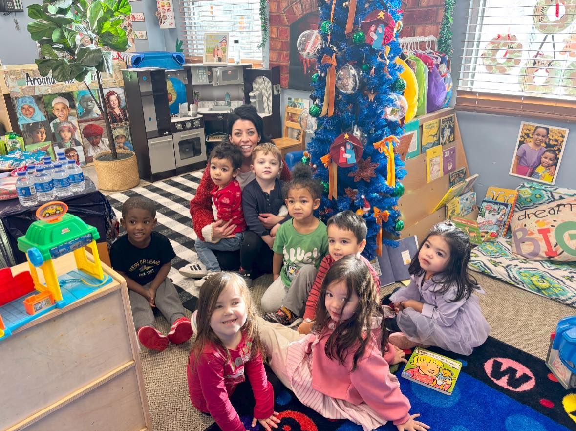 Children sitting on classroom carpet, smiling near a colorful holiday tree and toys.