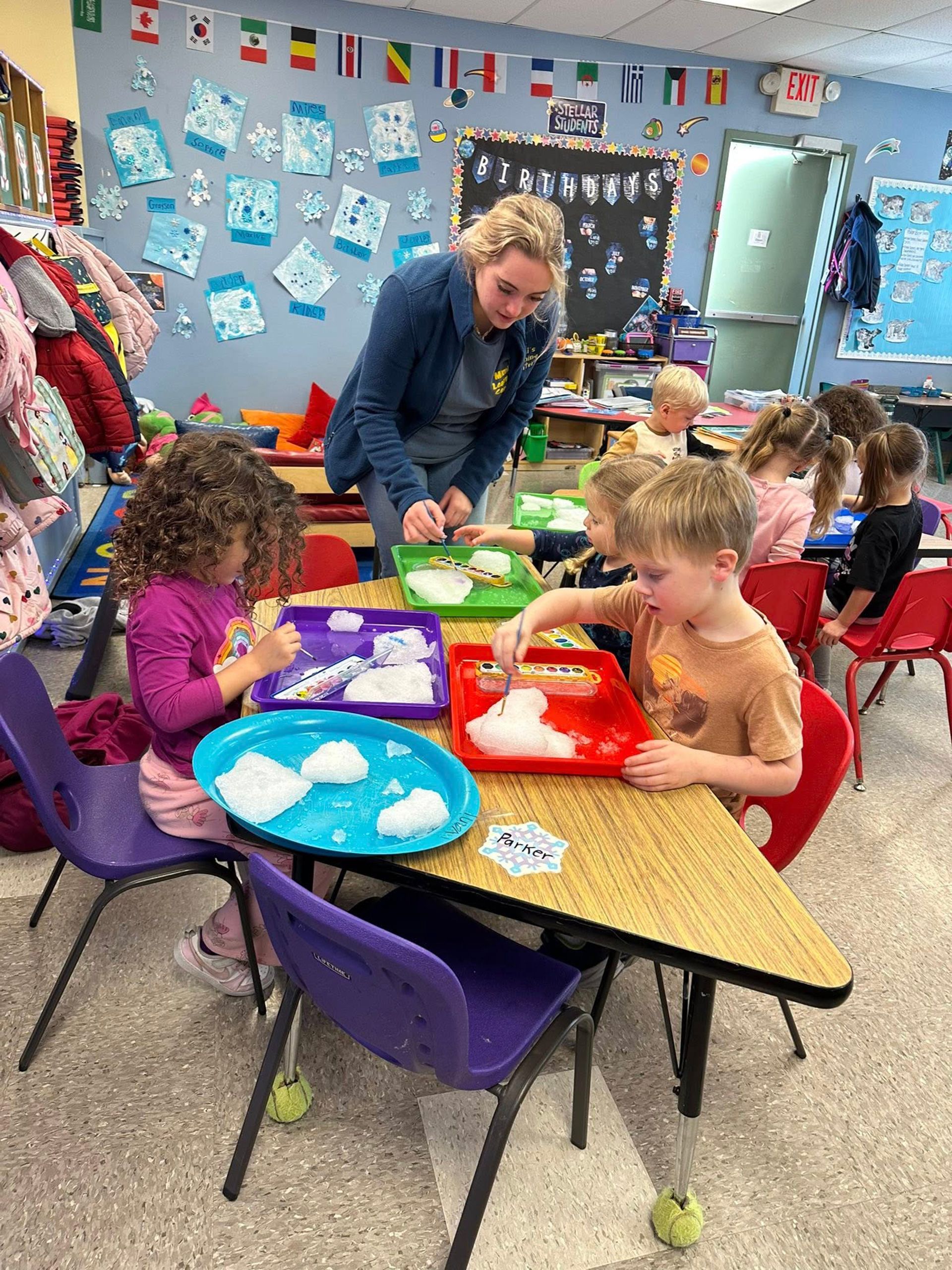 Children doing a tabletop activity with trays of white cotton-like material in a classroom, guided by a teacher.