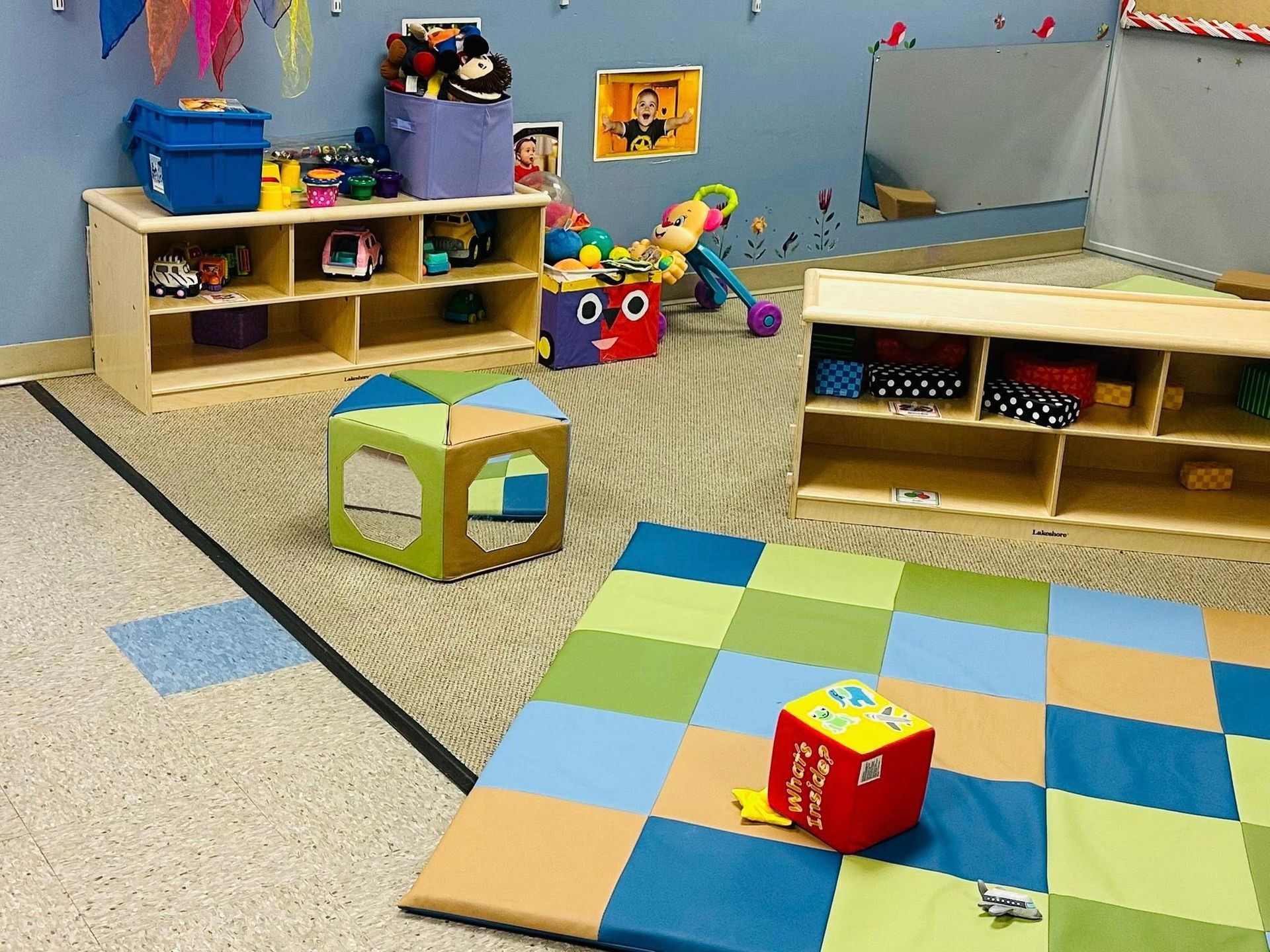 Colorful preschool playroom with shelves, toys, foam blocks, and a checkered mat on the floor