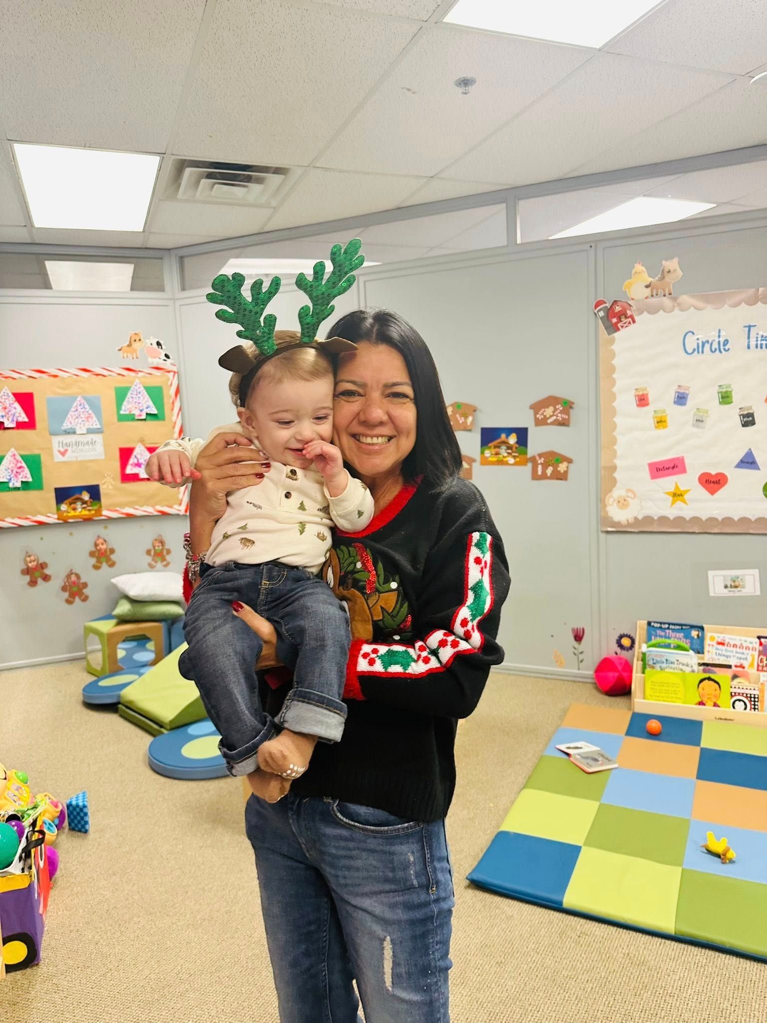 Adult holding child in a colorful classroom, both smiling with reindeer antlers during a holiday activity.