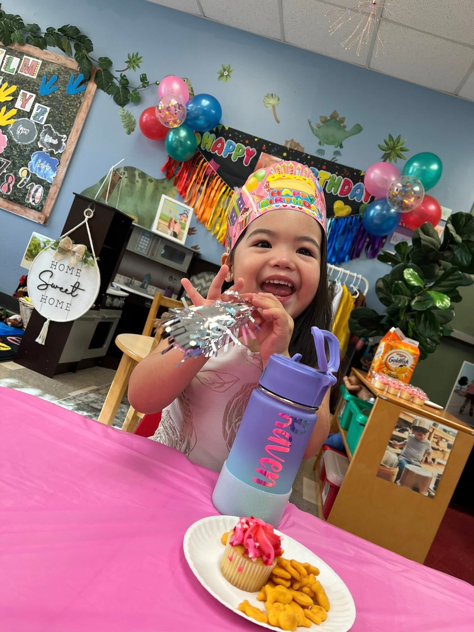 Smiling child at a pink party table with a cup and snack plate, surrounded by colorful balloons.
