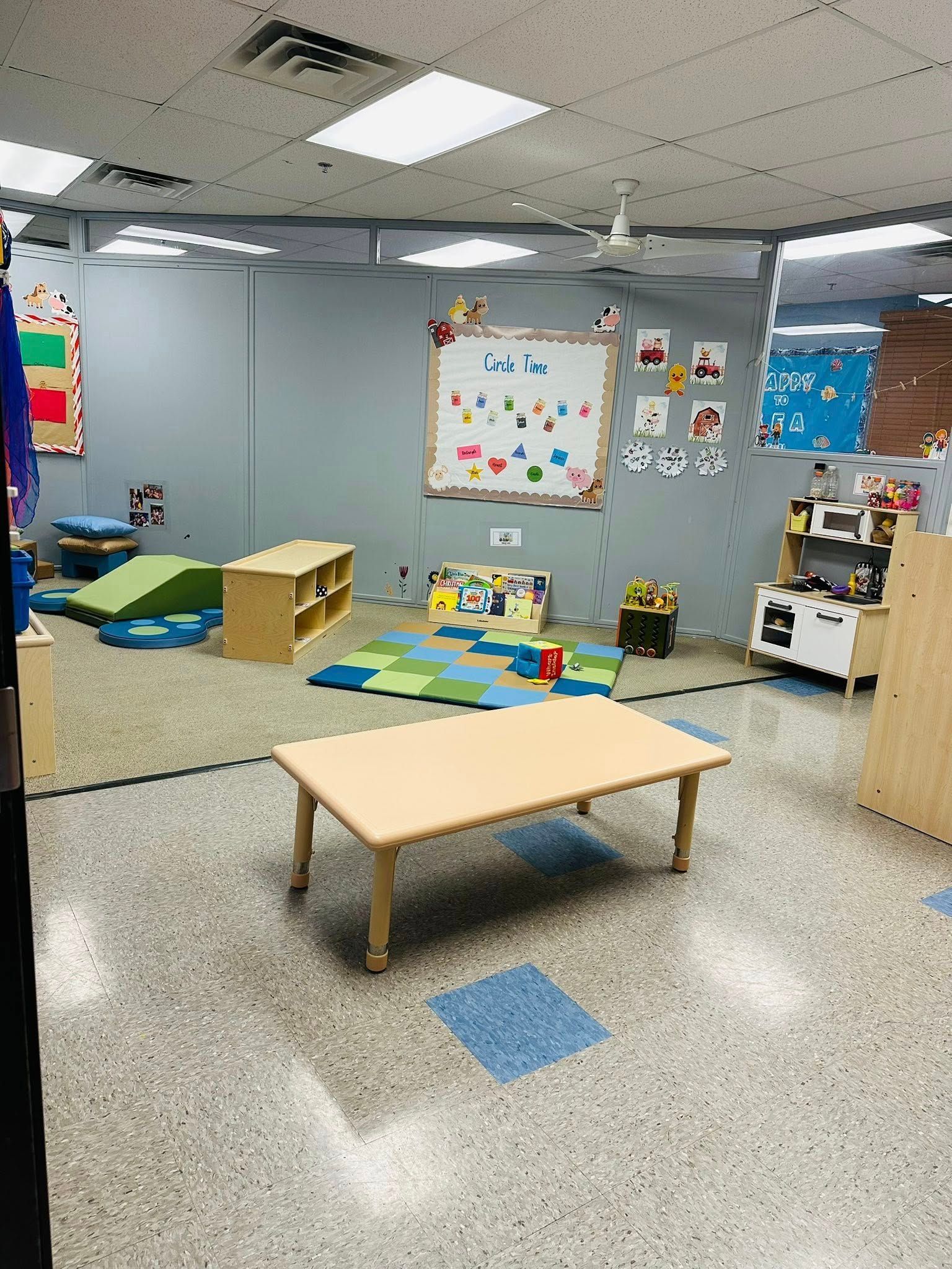 Colorful preschool classroom with low tables, rugs, wall art, and toys on a speckled floor.
