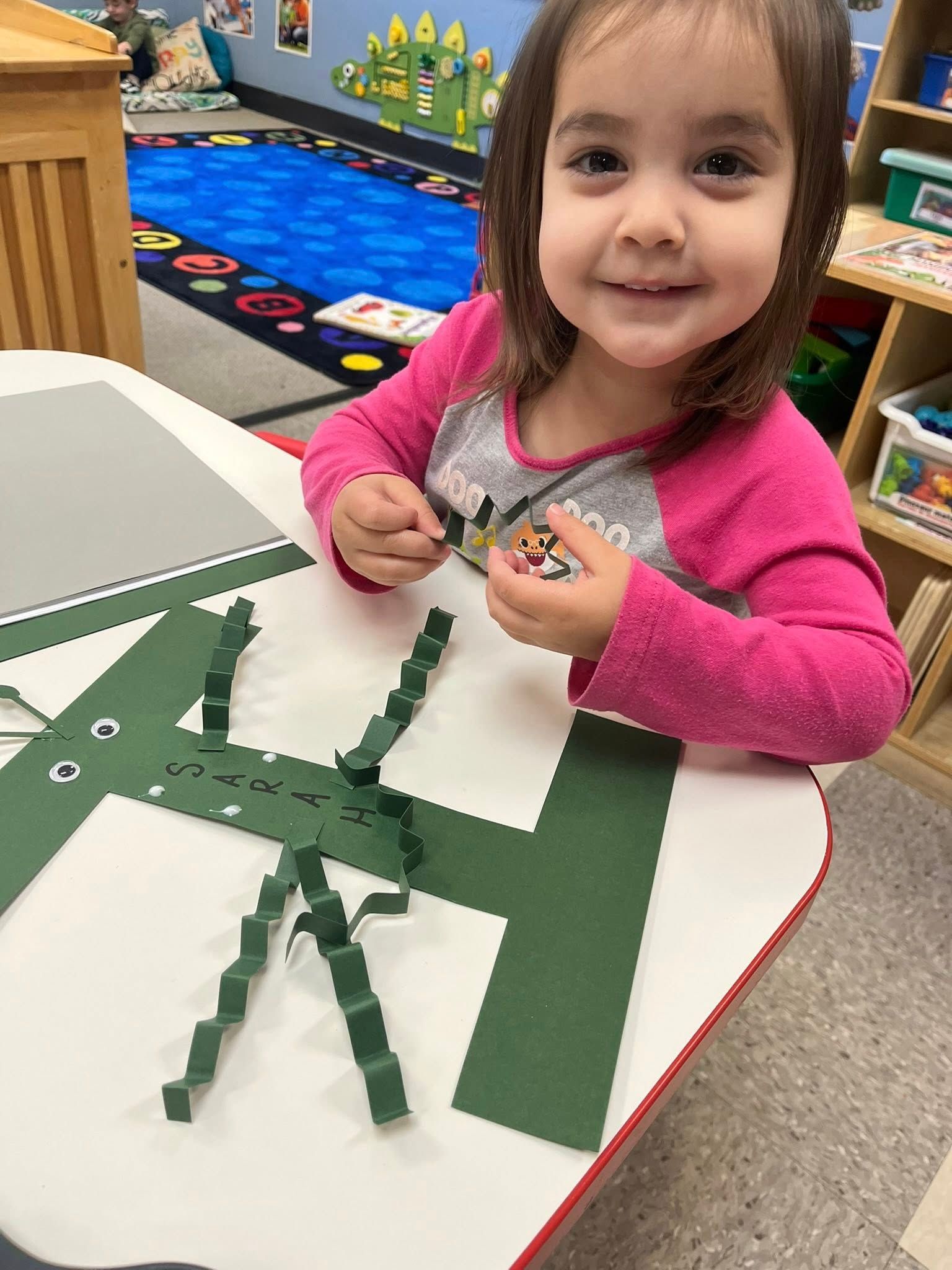 Child smiling at a table, building a green paper craft with strips in a classroom.