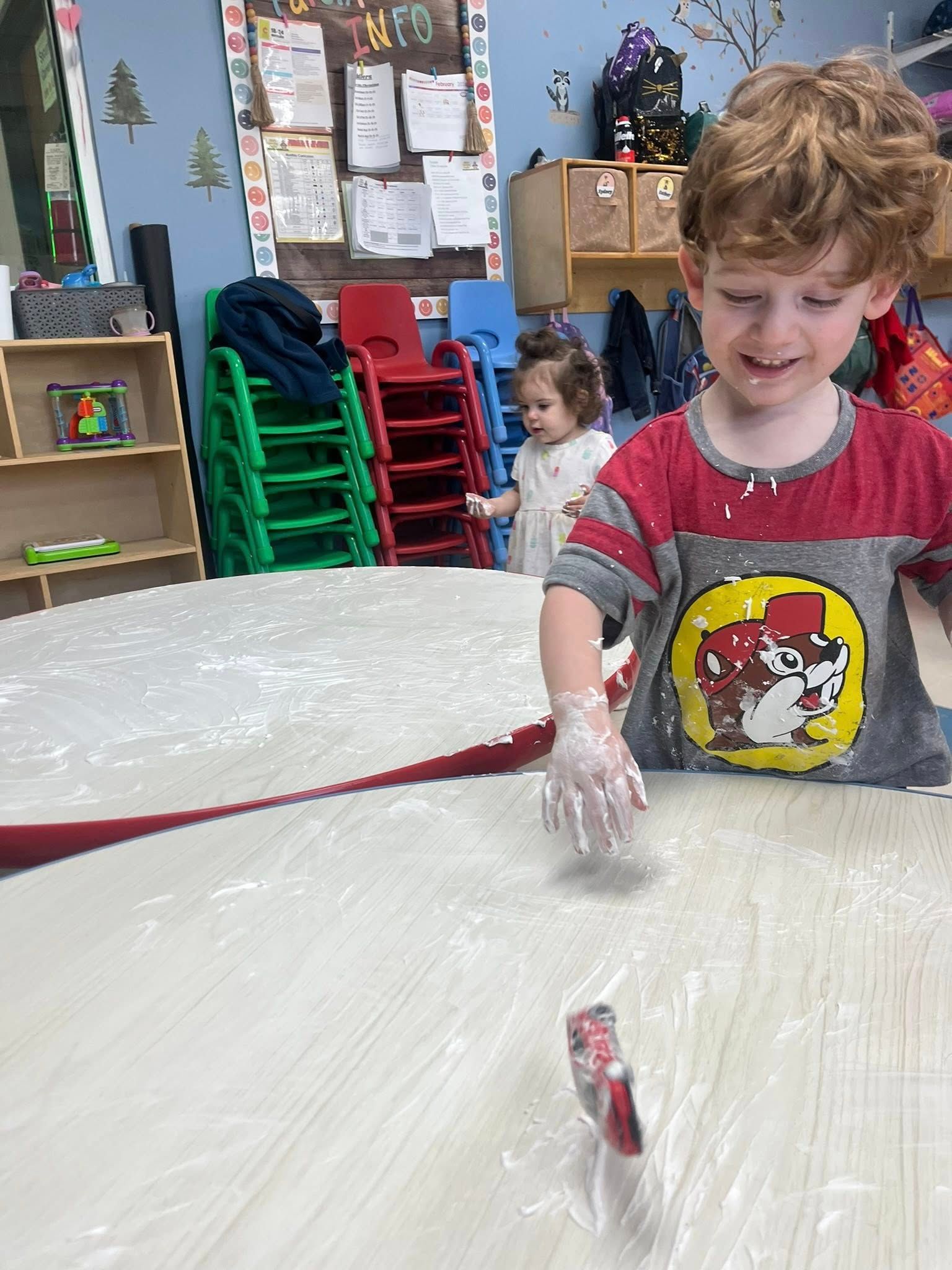 Child with messy white foam at a classroom table, smiling in a colorful preschool activity room.