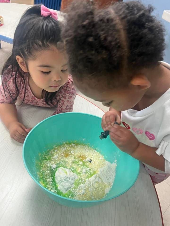 Two children leaning over a bowl, mixing ingredients together at a table