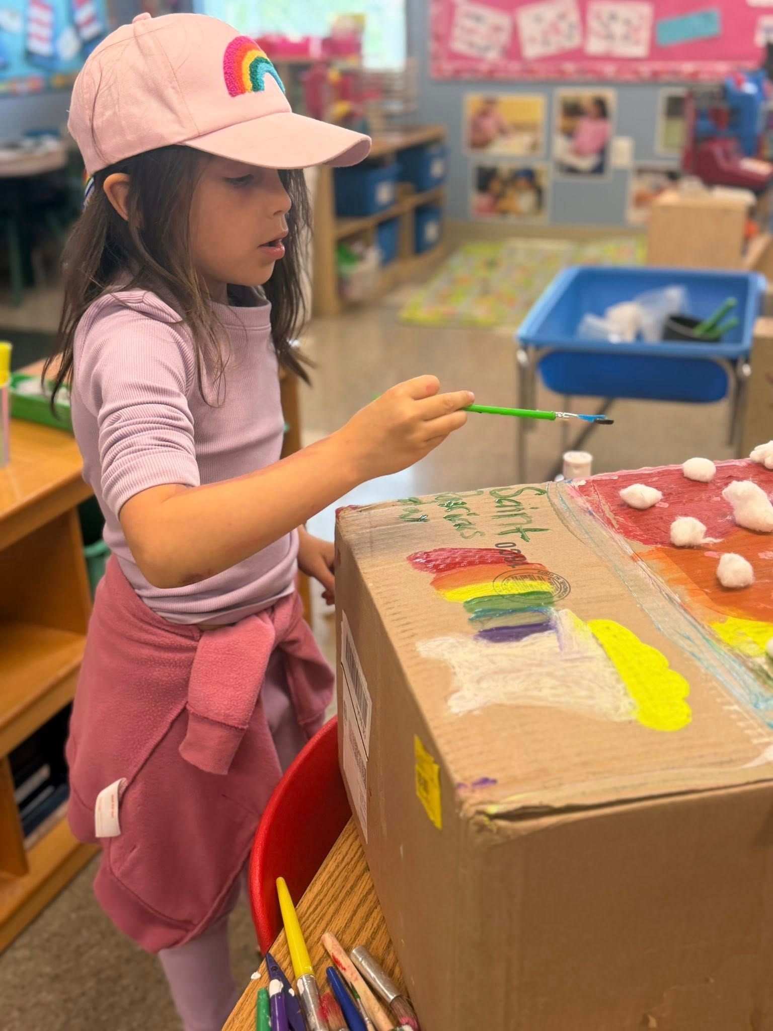 Child painting a cardboard box in a classroom, wearing a pink cap and pink clothes