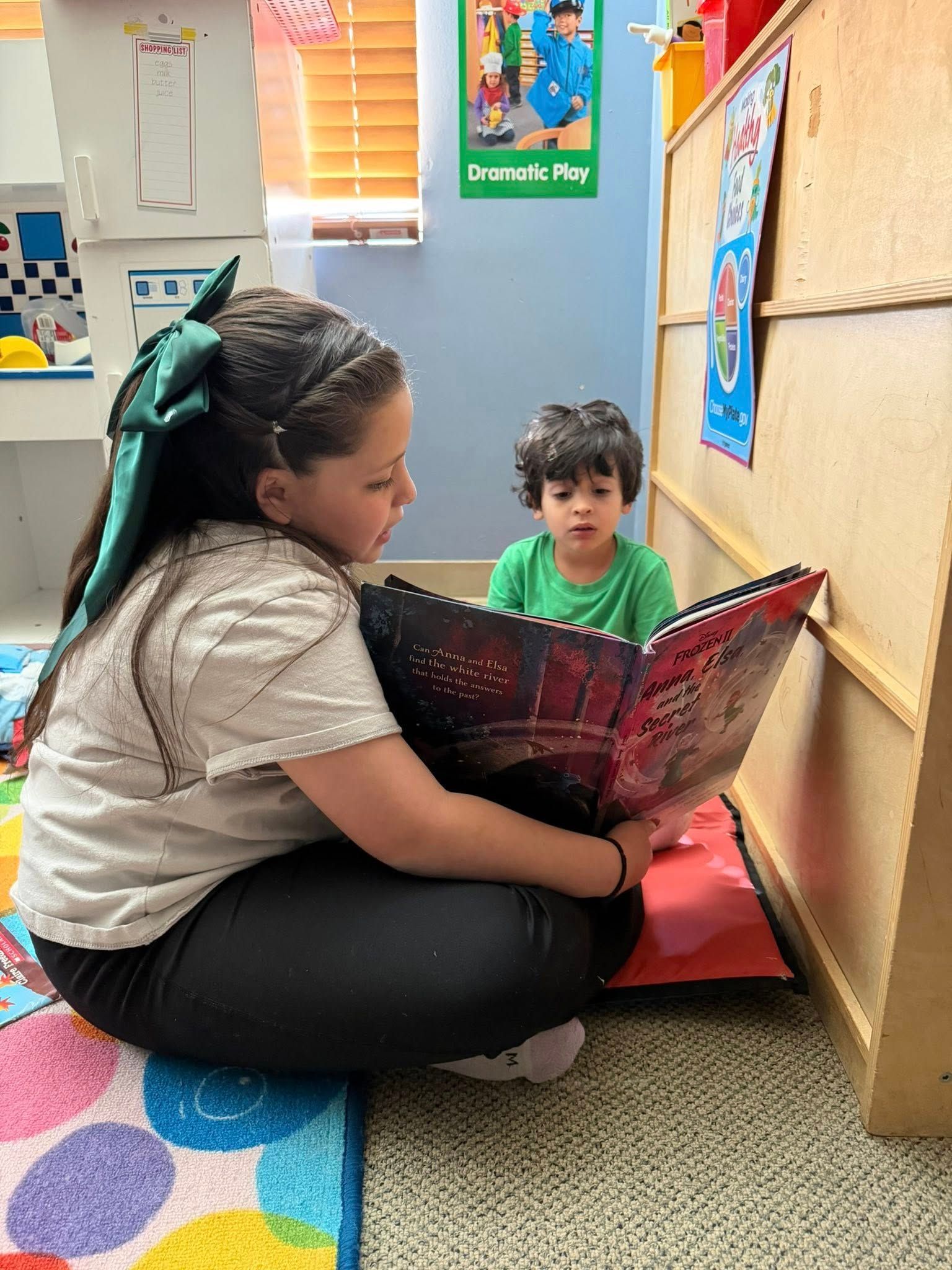 Child reading a book to a toddler in a colorful classroom corner