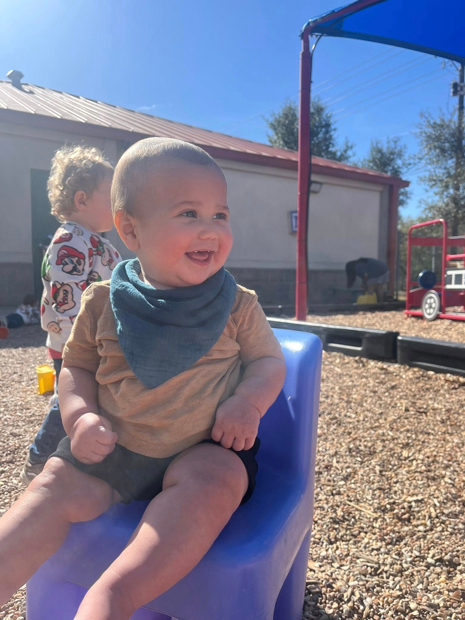 Smiling child in a blue seat outdoors on a sunny day, with another child and playground equipment behind.