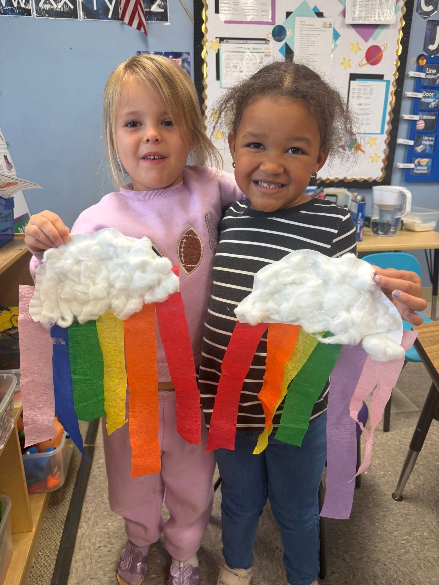 Two children holding colorful rainbow cloud crafts in a classroom, smiling at the camera.