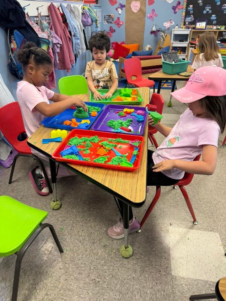 Children painting colorful trays at a classroom table with bright chairs and art supplies.