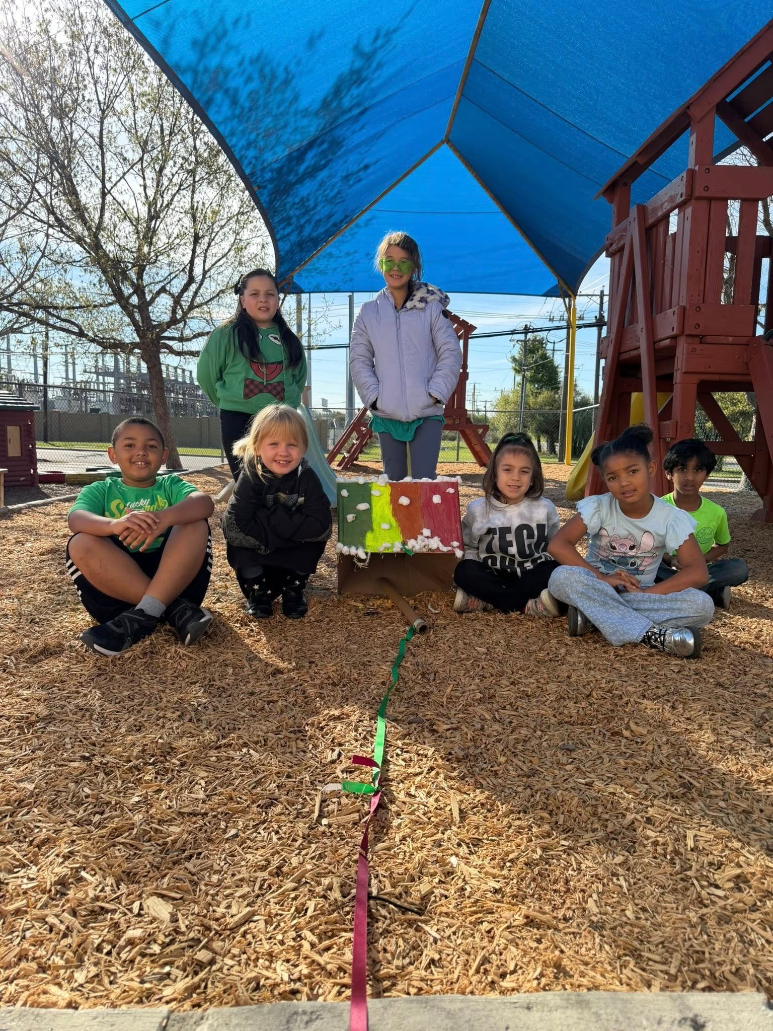 Children posing outdoors under a blue canopy, sitting and standing on wood chips near a playground structure.