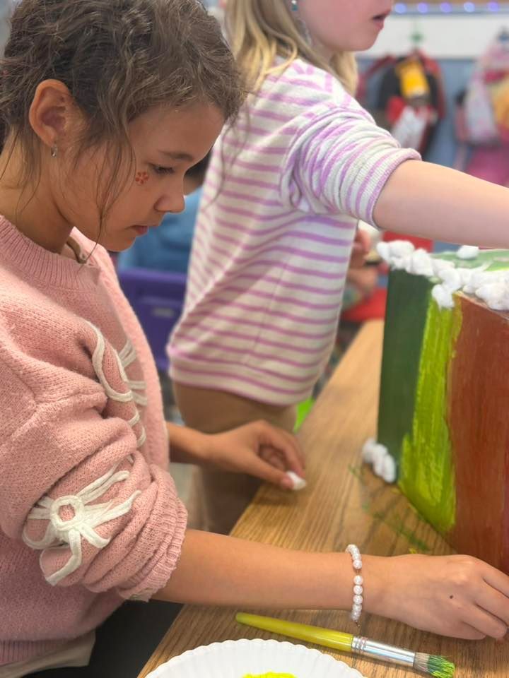 Children painting at a table, one in a pink sweater focused on a colorful craft project.