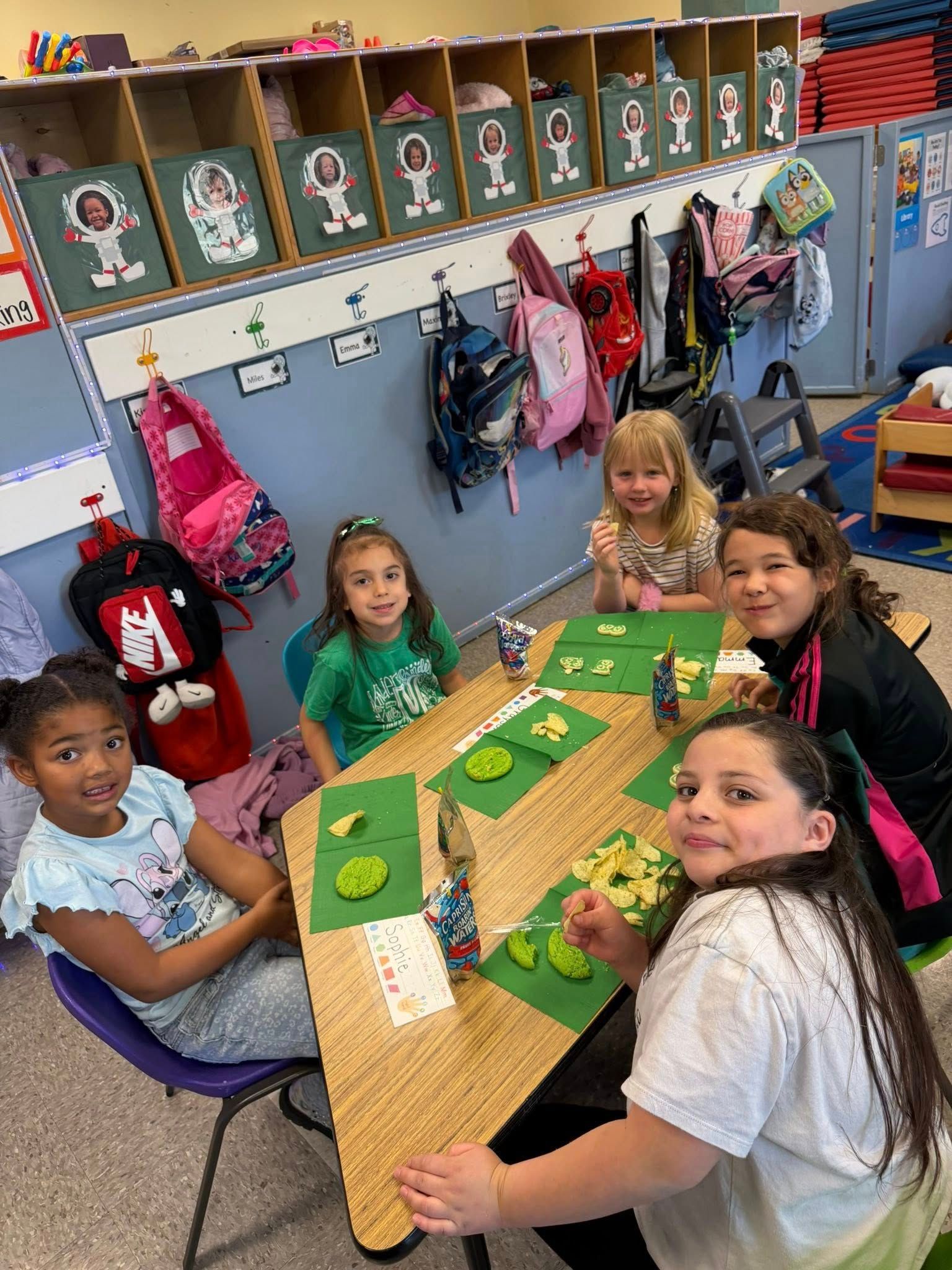 Children sit around a classroom table playing a board game, with backpacks and cubbies in the background.