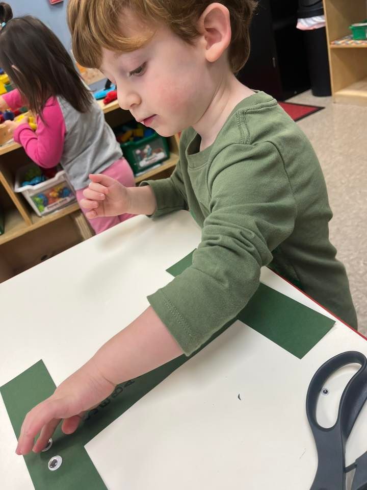 Child in a classroom glues green paper onto white cardstock beside scissors.