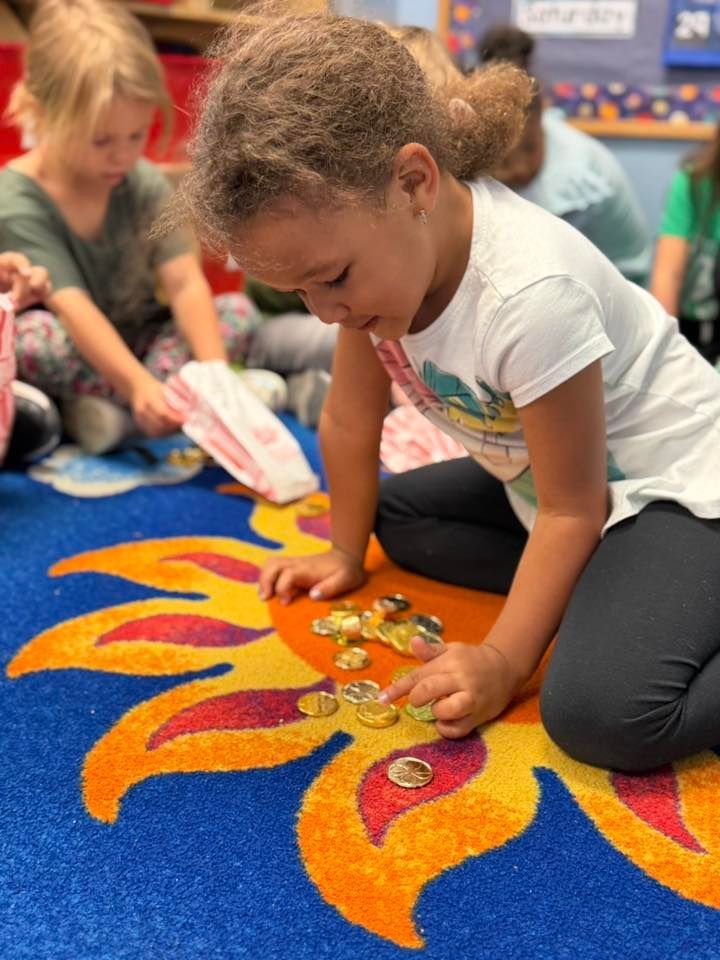 Child playing on colorful carpet with toy gems, crouched over a bright orange flame pattern in a classroom.
