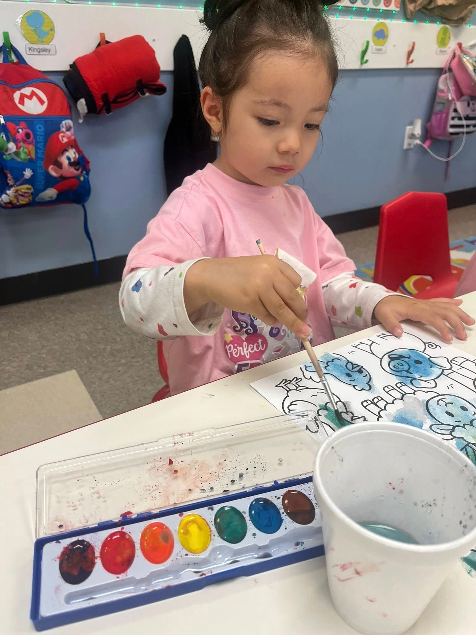 Child painting at a table with watercolor palette and cup, focused on a blue-and-white art project