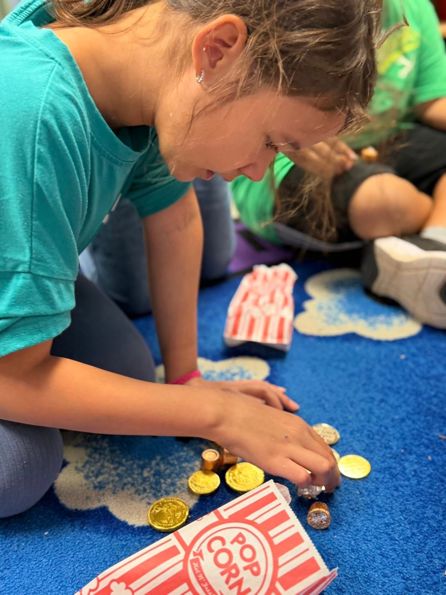 Child in a teal shirt leans over a blue carpet, sorting colorful pieces from a small popcorn tin.