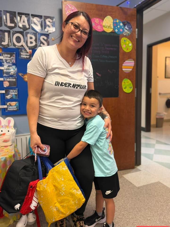 Woman smiling and hugging a child in a school hallway, holding colorful bags and standing by a bulletin board
