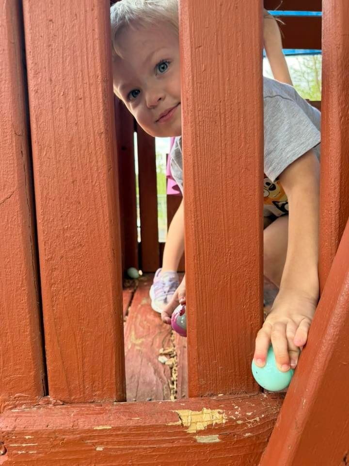Child peeking through wooden playset bars, holding a teal egg-shaped toy and looking at the camera.