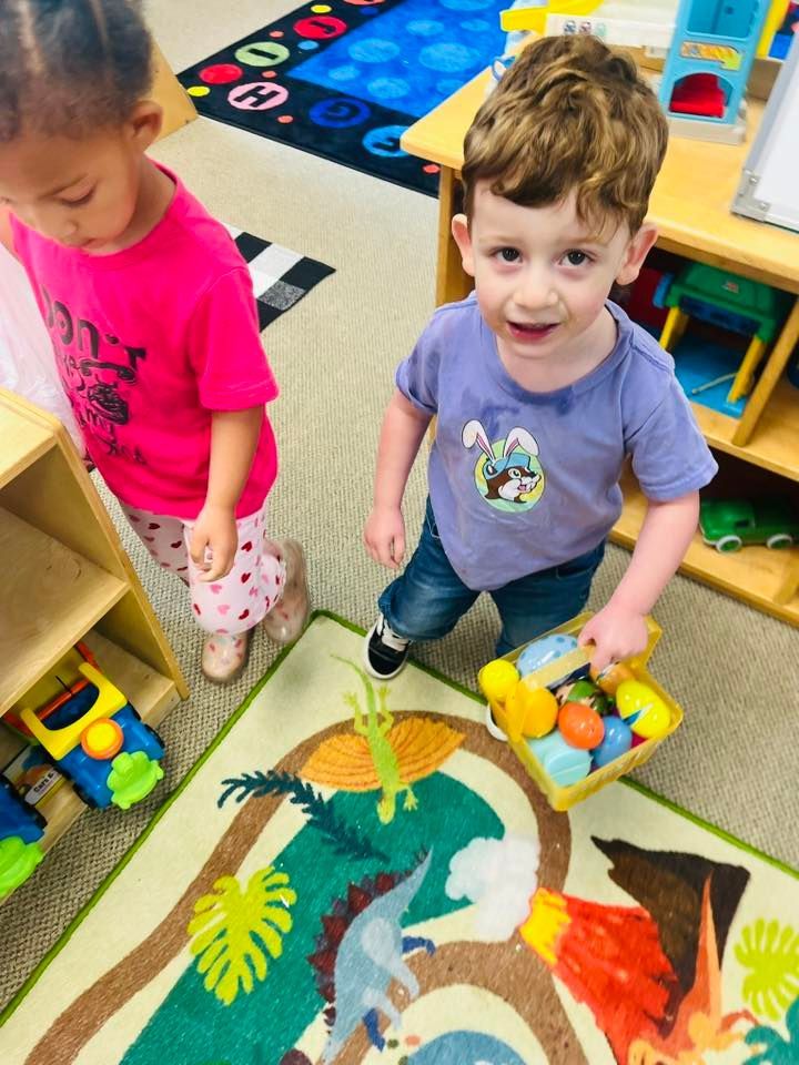 Children standing on a colorful classroom rug with animal illustrations and toys nearby