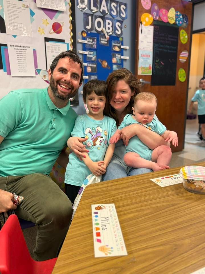 Smiling family of four seated at a classroom table with colorful wall displays in the background