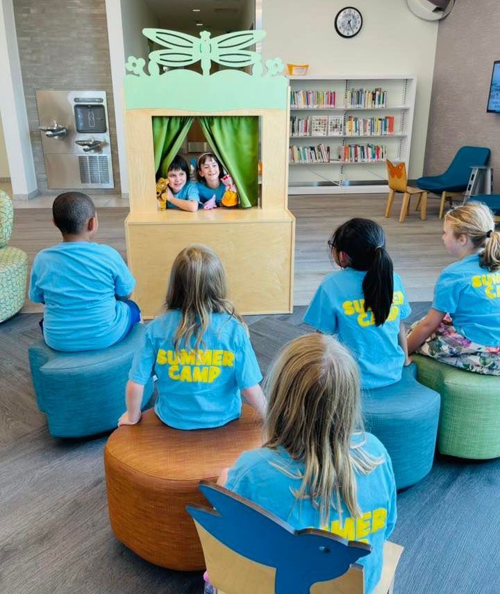 Children seated on colorful stools watch a puppet show in a bright library play area.