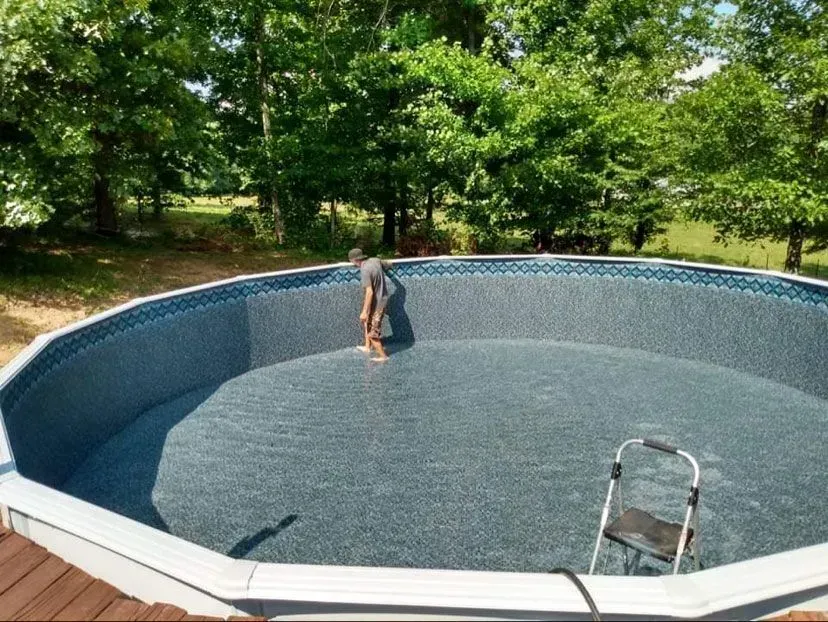 Man inside empty above-ground pool, cleaning floor. Trees in background, sunny day.