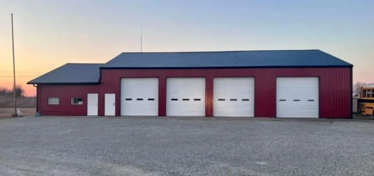 Red building with garage doors, small addition, blue roof, gravel parking. Evening sky.