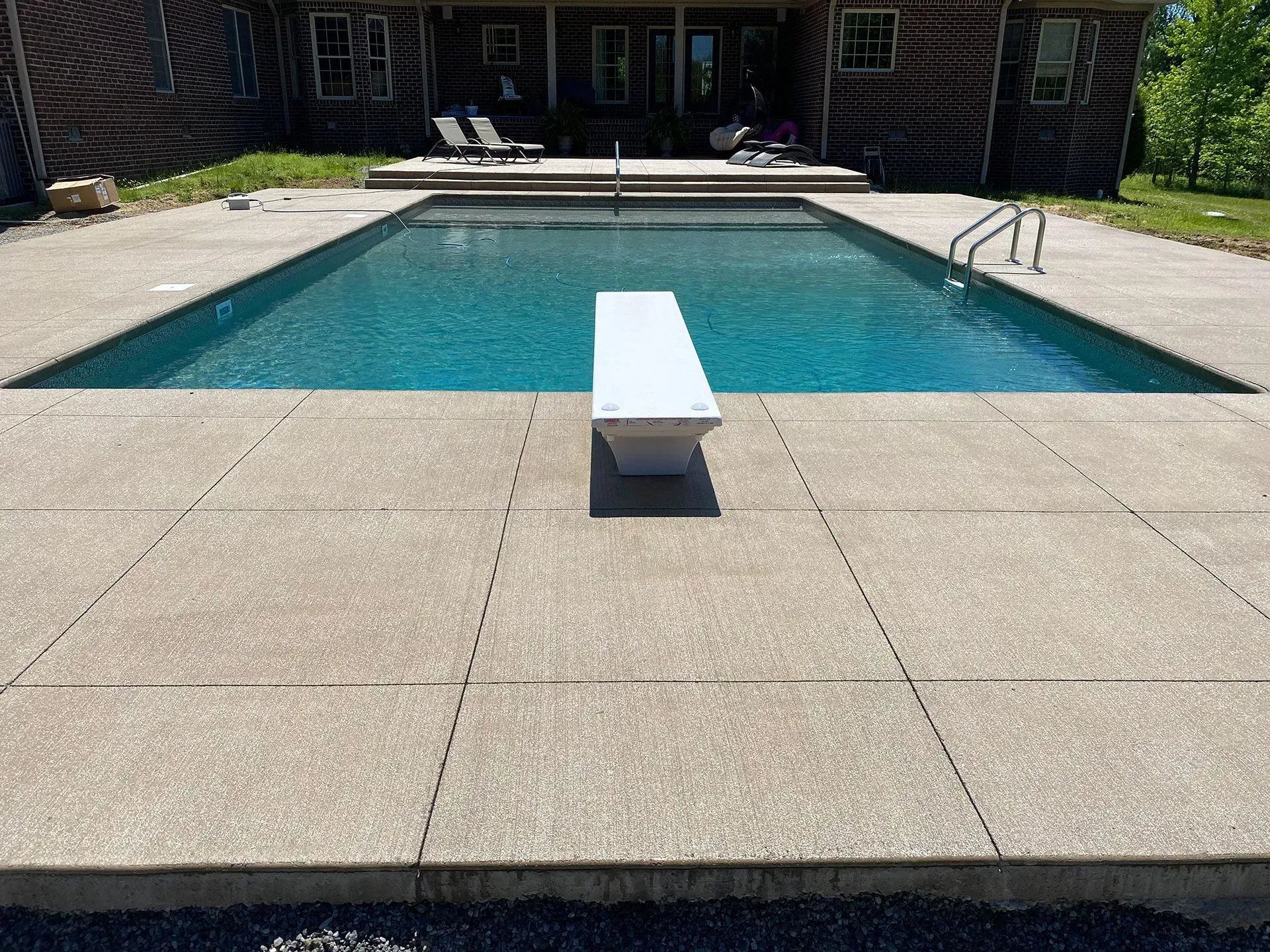 A rectangular swimming pool with a diving board.  Concrete patio surrounds the pool.  A building is in the background.