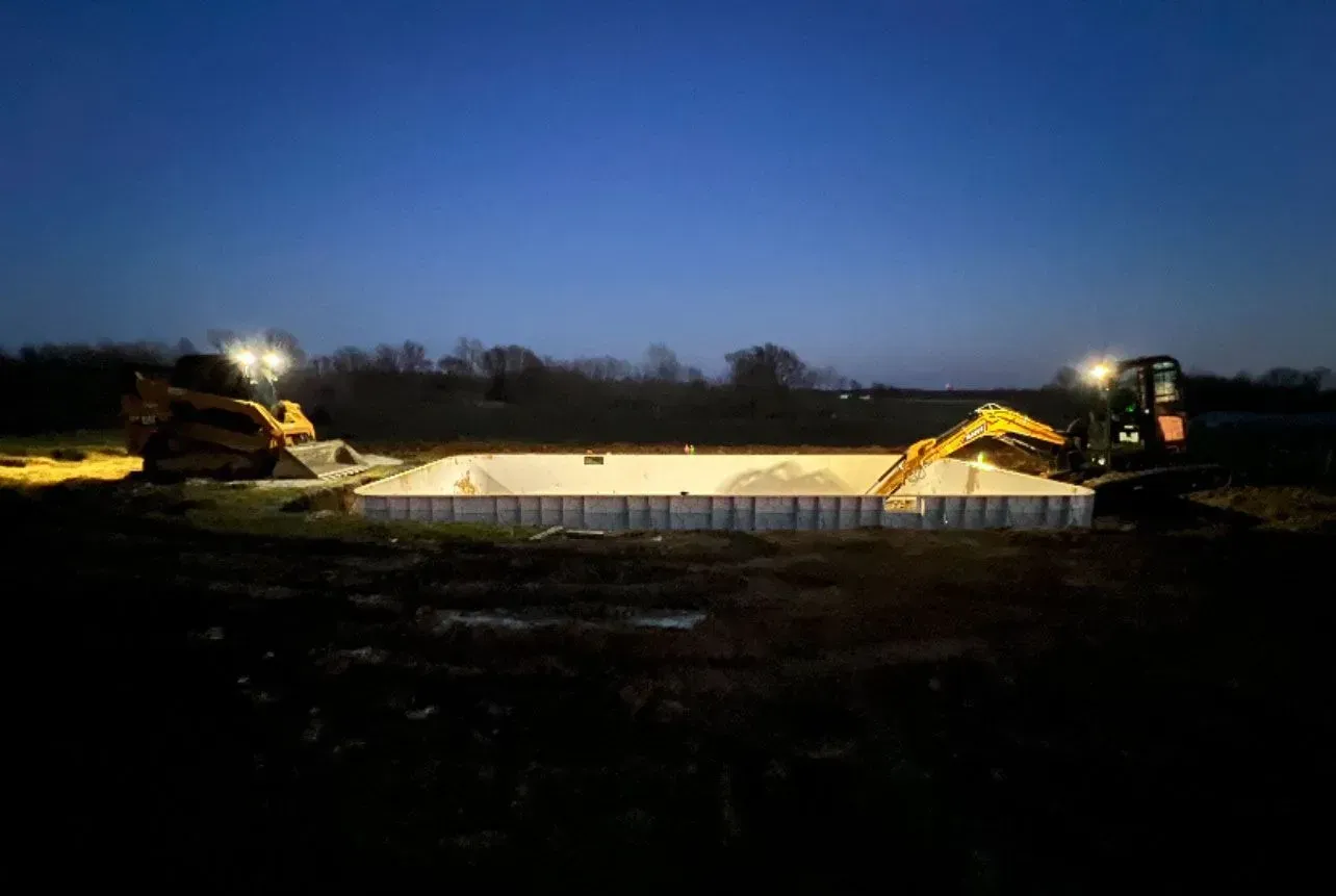 Construction site at night with excavators around an in-ground pool foundation, under a dark blue sky.